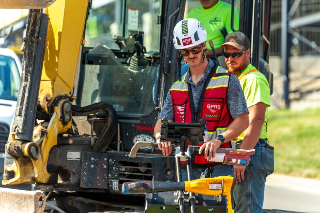 Construction workers watch a GPRS Project Manager using a ground penetrating radar utility locating cart.