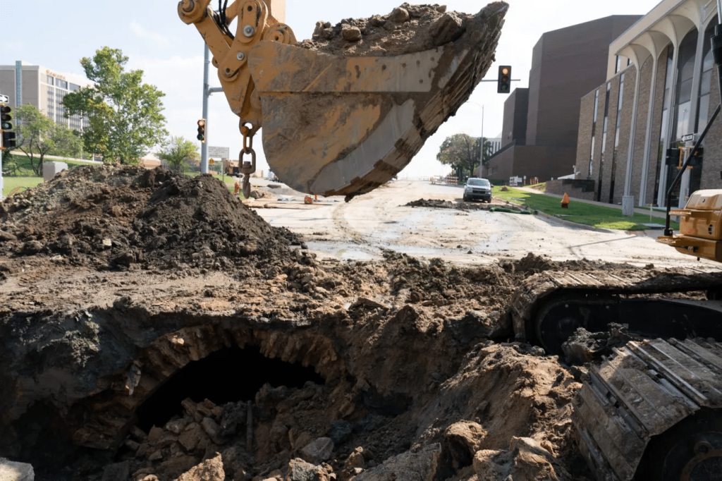 An excavator bucket pulling dirt out of a hole in the center of a road.