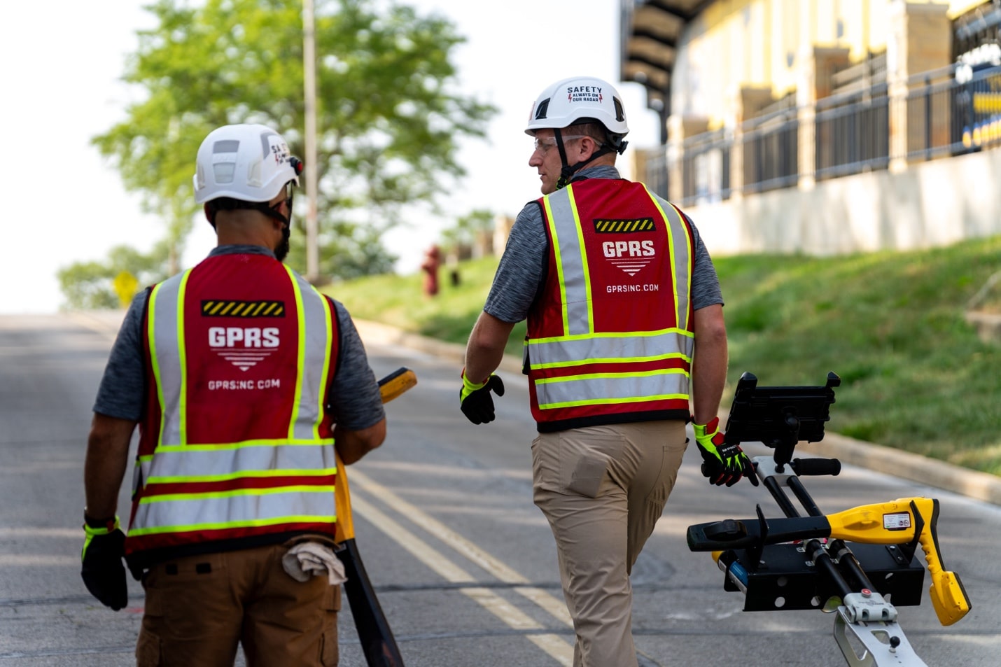 Two GPRS Project Managers carry ground penetrating radar scanning and electromagnetic locating equipment up a steep road.