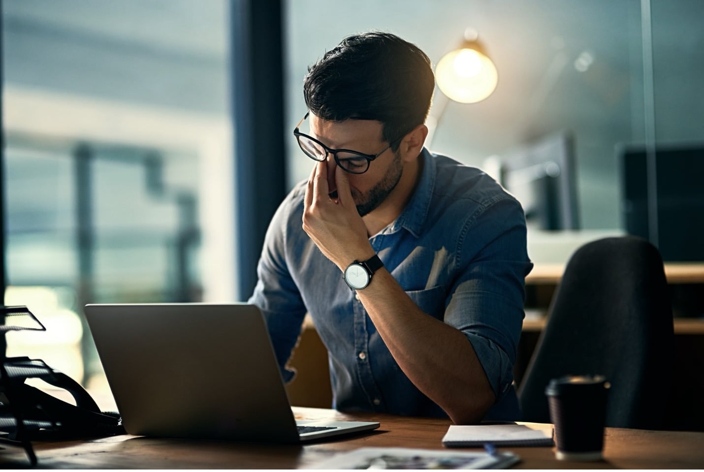 A man pinching his nose and pushing his glasses up his face while sitting in front of a laptop.