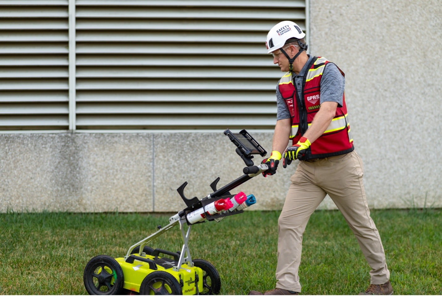 A GPRS Project Manager pushes a ground penetrating radar scanning cart along a grassy field.