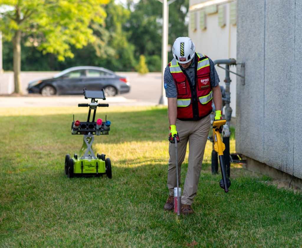 A GPRS Project Manager uses an electromagnetic locator and spray paint wand in a field next to a building, with a ground penetrating radar scanning cart in the background