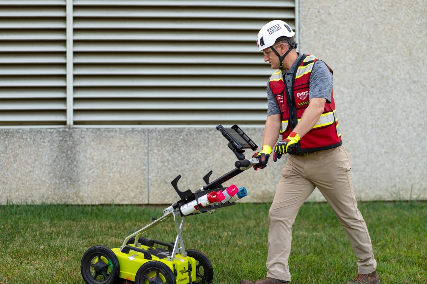 A GPRS Project Manager pushes a utility locating ground penetrating radar cart across a grass field.