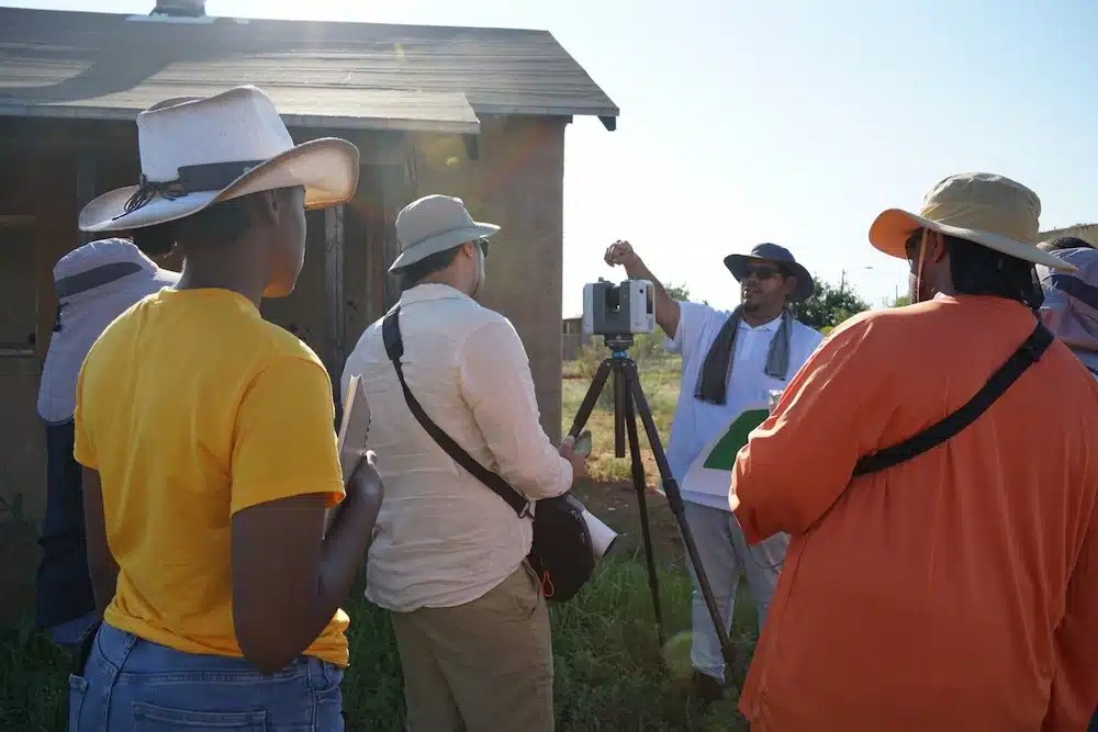 A group of students outdoors gathered around a Project Manager demonstrating how to use the Leica RTC360 laser scanner. A small building is visible in the background.