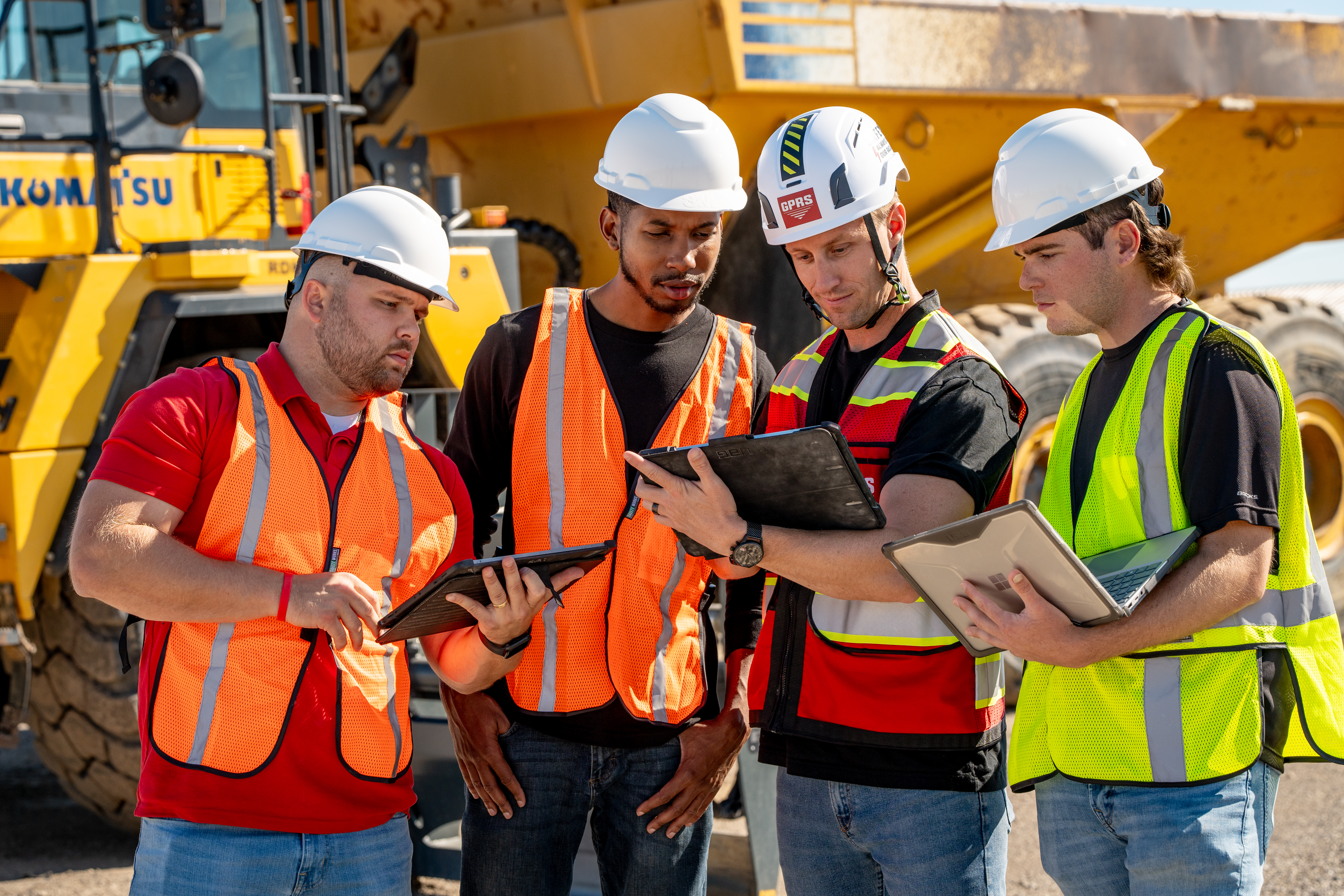 Four construction workers in safety vests and helmets review infrastructure records on tablets and laptops at a job site.