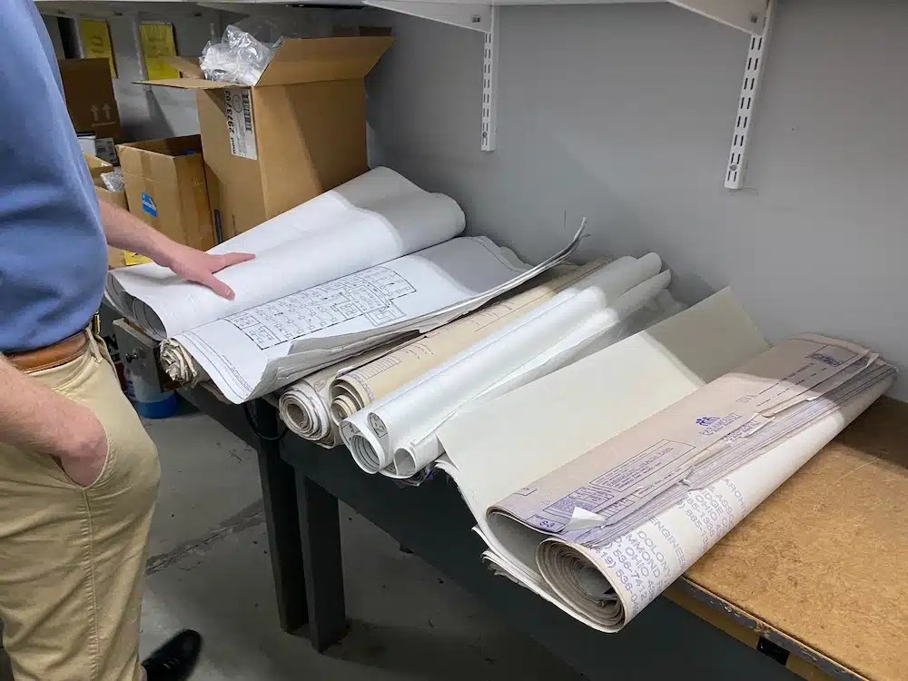 Rolled-up architectural blueprints and plans stored on a shelf with cardboard boxes, set against a concrete wall background.