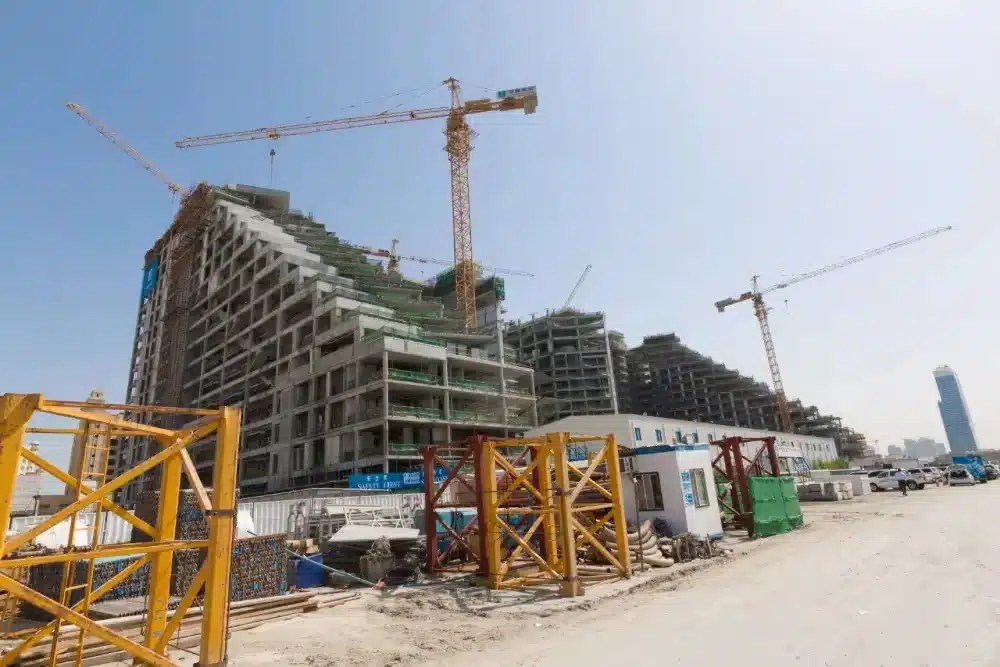 Construction site with multiple cranes and partially built, stepped-design buildings under a clear blue sky, with yellow and red metal structures and materials in the foreground.